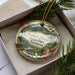 A decorative ornament with a landscape scene featuring mountains, pine trees, and a river, hanging from a string.