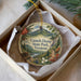 A decorative ornament in a box, featuring a scenic landscape with mountains, pine trees, and a river.