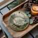 A decorative ornament featuring a lighthouse and a scenic landscape, placed on a wooden tray alongside pine cones and a lit candle.