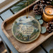 A decorative ornament featuring a lighthouse and a scenic view of Cape Meares State Scenic Viewpoint, Oregon, placed on a wooden tray alongside pine cones and a lit candle.