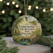 A round ornament with a scenic landscape featuring a river, trees, and a bridge, hanging in front of a blurred background with twinkling lights.