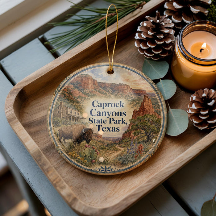 A decorative ornament featuring a scenic image of Caprock Canyon State Park, Texas, with a bison and desert landscape. The ornament is placed on a wooden tray, accompanied by pine cones and a lit candle.
