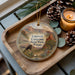 A decorative ornament featuring a scenic image of Caprock Canyon State Park, Texas, with a bison and desert landscape. The ornament is placed on a wooden tray, accompanied by pine cones and a lit candle.