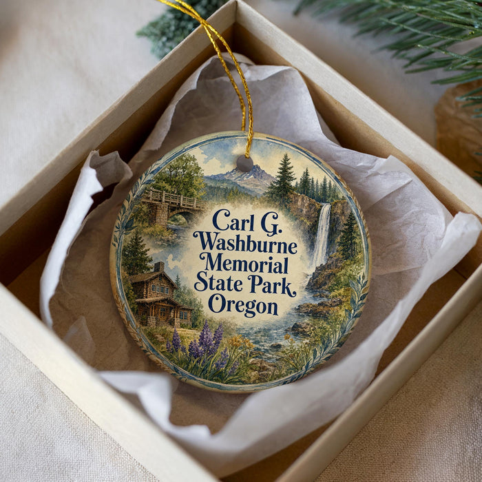 A decorative ornament in a white box, featuring a scenic landscape with a waterfall, cabin, and mountains.