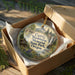A round ornament in a brown box, featuring a scenic landscape with a mountain, trees, and a waterfall.