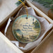 A round ornament with a lighthouse and beach scene, placed in a box with a white tissue paper lining.