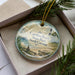 A decorative ornament in a box, featuring a lighthouse and a beach scene, with the text "Carolina Beach State Park North Carolina" printed on it.