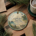 A round ornament with a lighthouse and beach scene hangs on a wooden surface, surrounded by pine branches and a cup of coffee.