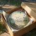 A decorative ornament in a wooden box, featuring a lighthouse and a scenic landscape, with the text "Carolina Beach State Park, North Carolina" printed on it.