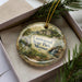 A decorative ornament in a box, featuring a scenic landscape with a bridge, trees, and water.