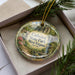 A decorative ornament in a box, featuring a scenic landscape with a waterfall and greenery, with the text "Cathedral State Park, West Virginia" prominently displayed.