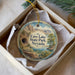 A decorative ornament in a box, featuring a scenic landscape with mountains, trees, and a body of water.