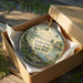 A decorative ornament in a box, featuring a lighthouse and a scenic landscape, with the text "Cedar Point State Park New York" printed on it.