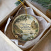 A decorative ornament in a box, featuring a scenic image of a historical site with a castle, boats, and lush greenery.