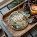 A wooden tray with a decorative ornament featuring a scene of a river, boats, and a building, along with pine cones and a lit candle.