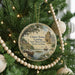 A round ornament hanging on a Christmas tree, featuring a scenic image of a town with a river and palm trees.