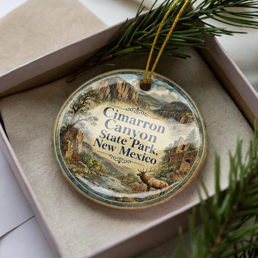 A decorative ornament with a landscape scene featuring a river, mountains, and a deer, placed in a box with pine branches.