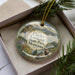 A decorative ornament in a box, featuring a scenic landscape with mountains, pine trees, and a castle-like structure.