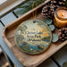 A decorative ornament with a scenic lake and park scene, featuring a wooden tray with pine cones and a lit candle.