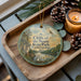 A wooden tray with a decorative ornament featuring a scenic landscape of cliffs, trees, and a river, along with pine cones and a lit candle.