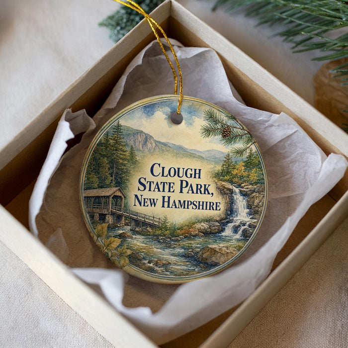 A decorative ornament in a box, featuring a scenic image of Clough State Park in New Hampshire with a waterfall and a wooden bridge.