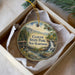 A decorative ornament in a box, featuring a scenic image of Clough State Park in New Hampshire with a waterfall and a wooden bridge.