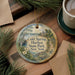 A decorative ornament with a scenic image of Cold Spring Harbor State Park, New York, hangs on a wooden surface alongside a cup of coffee and some pine branches.