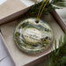 A decorative ornament in a box, featuring a scenic landscape with mountains, waterfalls, and greenery.
