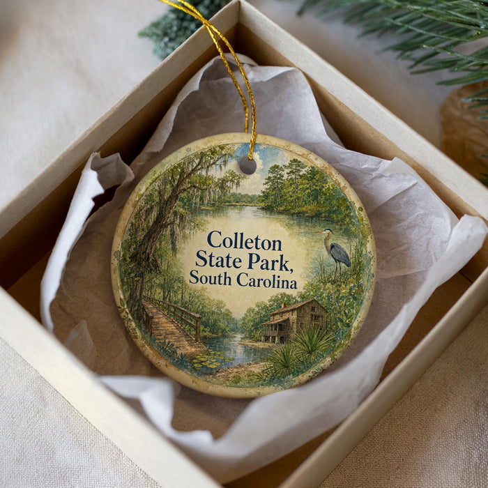 A round ornament in a box, featuring a scenic landscape with a bridge, trees, and a bird, with the text "Colleton State Park, South Carolina" printed on it.