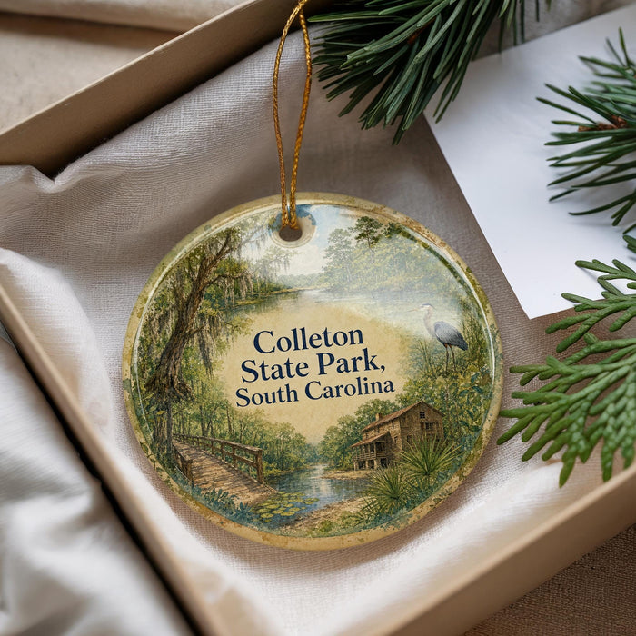 A round ornament with a painting of a park scene, featuring a house, trees, and a bird, is displayed in a box with pine branches.