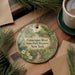 A decorative ornament with a scenic image of the Connecticut River and the State Park Preserve in New York, featuring a bird and a deer, is displayed on a wooden surface alongside a cup of coffee and some pine branches.