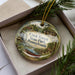 A decorative ornament in a box, featuring a scenic landscape with a river, mountains, and pine trees.