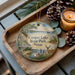 A wooden tray with a decorative ornament featuring a scenic landscape of Cooper Lake State Park, Texas, along with pine cones and a lit candle.