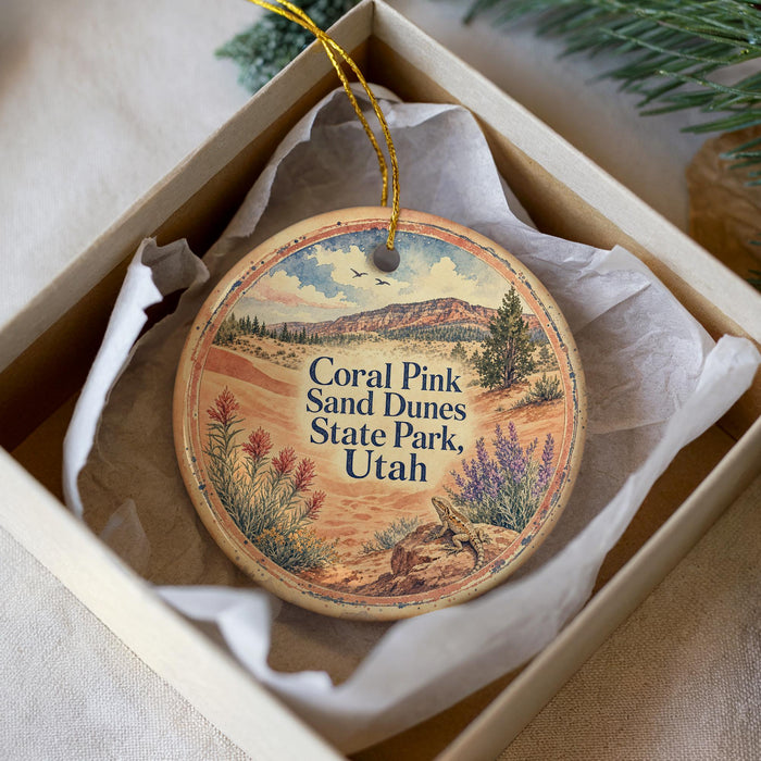 A decorative ornament in a box, featuring a landscape with mountains, trees, and a bird, along with the text "Coral Pink Sand Dunes State Park, Utah".