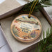 A decorative ornament with a landscape scene featuring a mountain, desert, and various plants, placed in a box with pine branches.