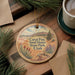 A decorative ornament featuring a scenic landscape with mountains, trees, and a desert setting, hanging on a wooden surface.