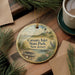 A decorative ornament with a lighthouse and a boat, hanging on a wooden surface, surrounded by pine branches and a cup of coffee.