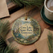 A decorative ornament with a scenic landscape of Cove Lake State Park, Tennessee, featuring a bird, flowers, and a cabin, is displayed on a wooden surface alongside a cup of coffee and some pine branches.