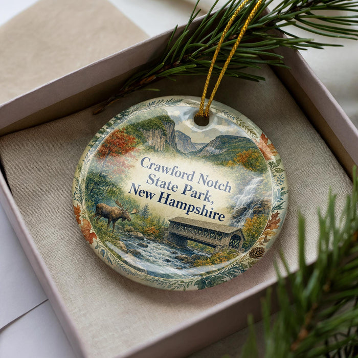 A decorative ornament in a box, featuring a scenic landscape with a river, mountains, and a bridge.