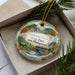 A decorative ornament in a box, featuring a scenic landscape with a river, mountains, and a bridge.