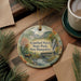 A decorative ornament featuring a scenic image of Crawford Notch State Park in New Hampshire, with a moose and a bridge in the background. The ornament is hanging from a string and is placed on a wooden surface, surrounded by pine branches and a cup of coffee.