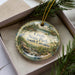 A decorative ornament in a box, featuring a scenic landscape with a river, trees, and a bird.