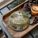 A decorative ornament featuring a scenic image of Croft State Park, South Carolina, with pine cones and a lit candle on a wooden tray.