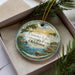 A decorative ornament in a box, featuring a scenic landscape with a lake, trees, and mountains.