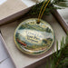 A decorative ornament in a box, featuring a scenic landscape with a lake, mountains, and a canoe, along with the text "Crystal Lake State Park, Vermont".