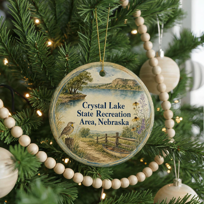 A round ornament hanging on a Christmas tree, featuring a scenic landscape with a lake, mountains, and a bird.