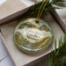 A decorative ornament in a box, featuring a scenic landscape with a river, bridge, and wildlife.