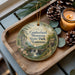 A decorative ornament featuring a scenic landscape of Cumberland Mountain State Park, Tennessee, with a waterfall and pine trees. The ornament is placed on a wooden tray, accompanied by a lit candle and pine cones.