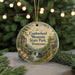 A round ornament with a scenic image of a waterfall and mountains, surrounded by greenery, hangs in front of a blurred Christmas tree with lights.