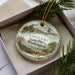 A decorative ornament in a box, featuring a scenic landscape with mountains, pine trees, and a bird in flight.